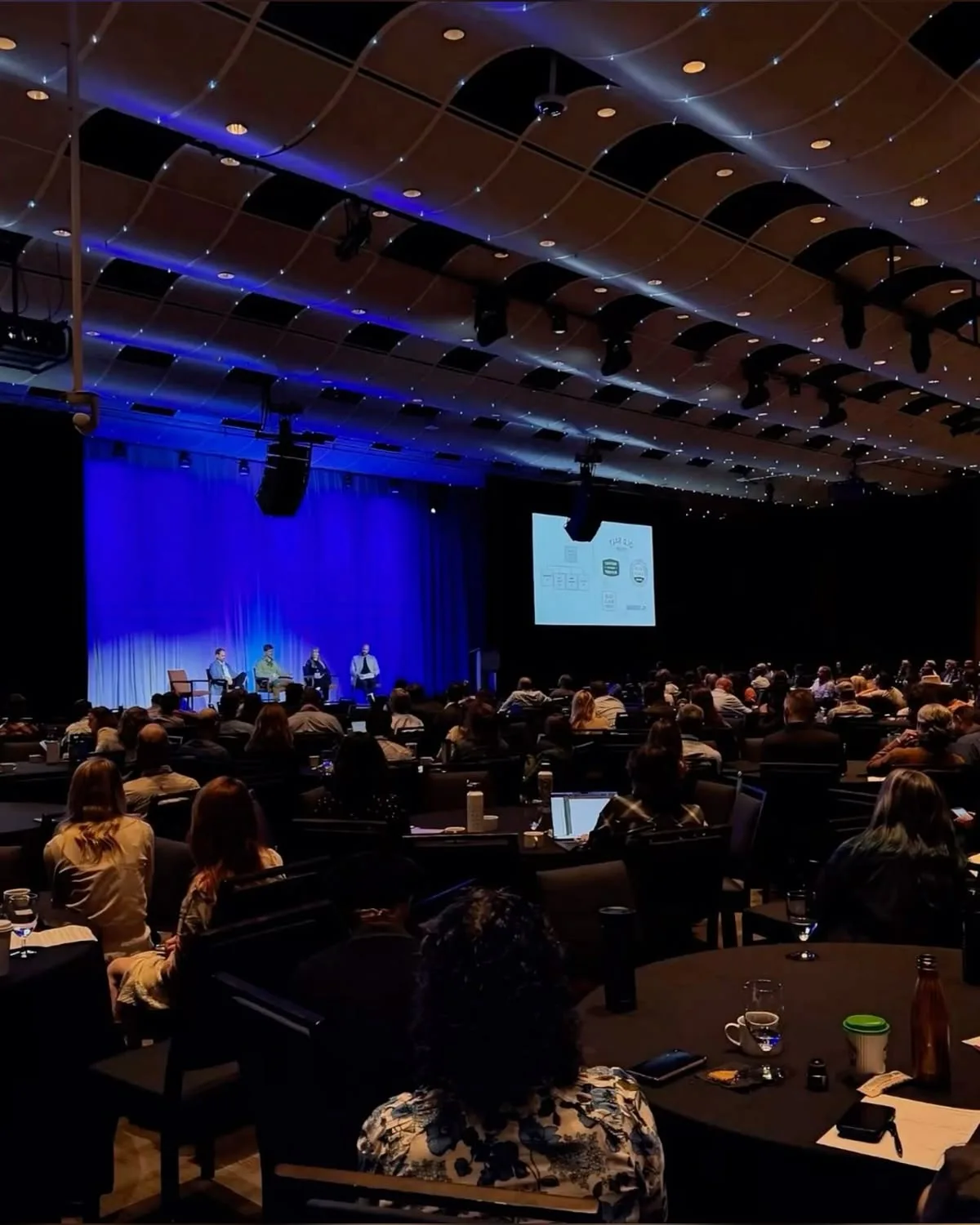 A conference audience listens to a panel on a lit stage.