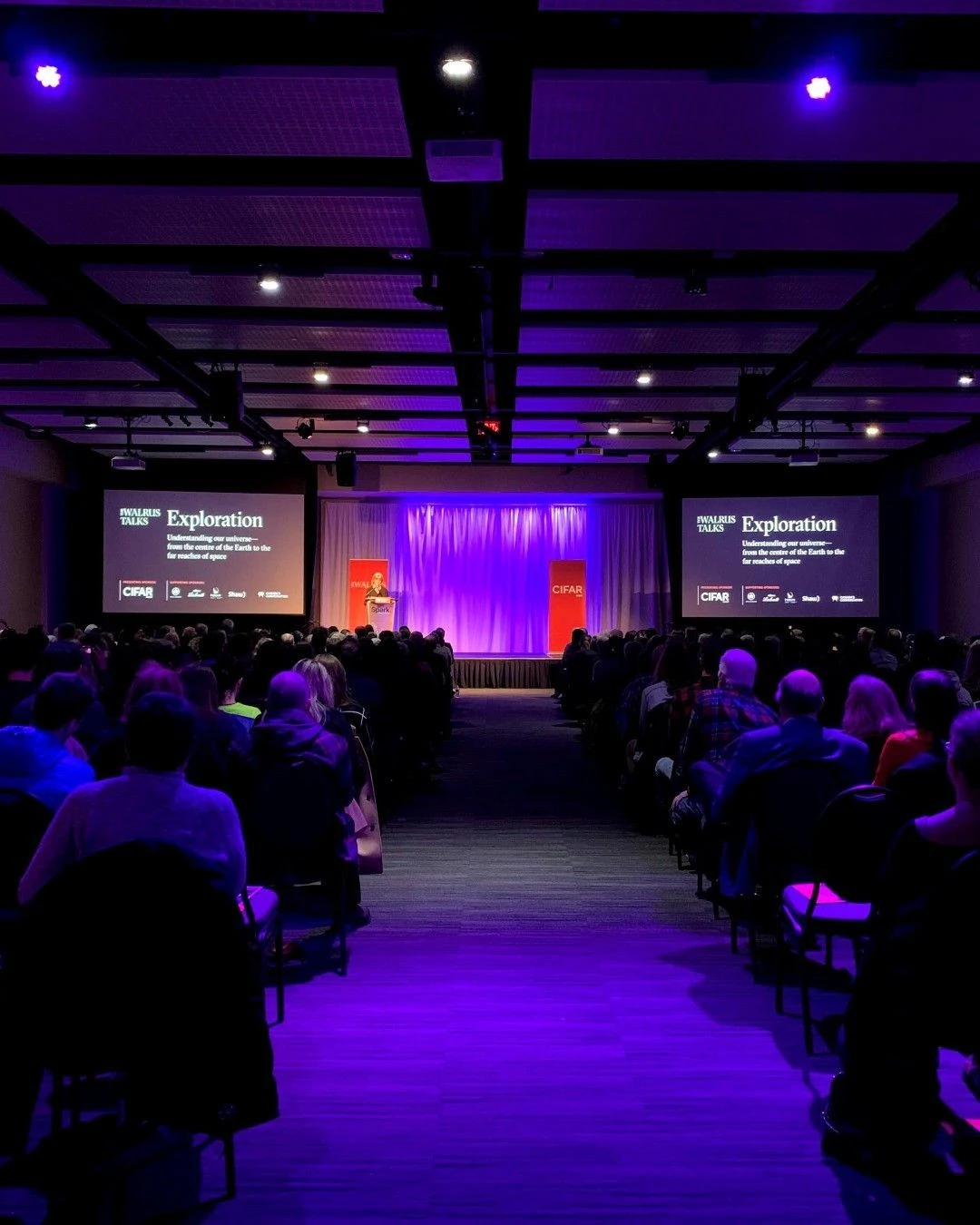 An audience faces an illuminated conference stage.